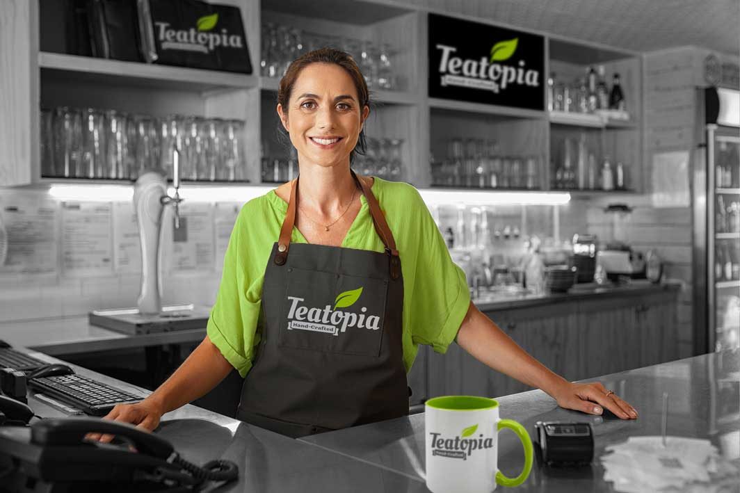 A café owner behind her counter, with the Teatopia logo on her apron, mug and signs
